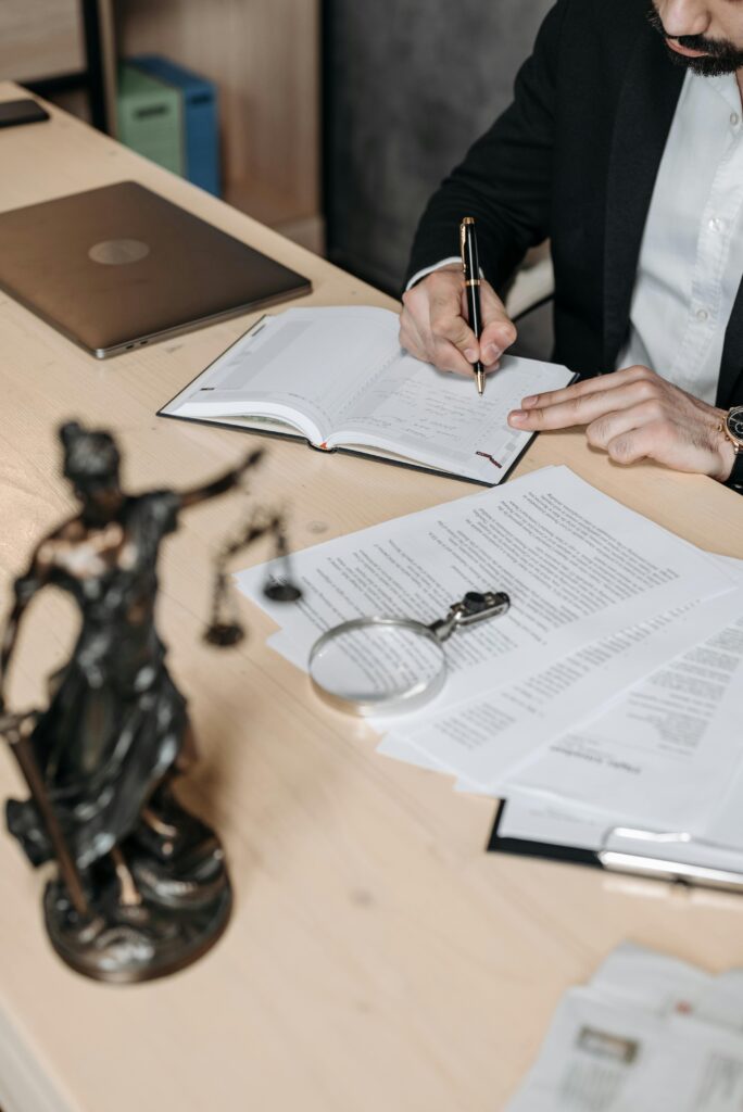 Professional working on documents at desk with legal statue and magnifying glass.